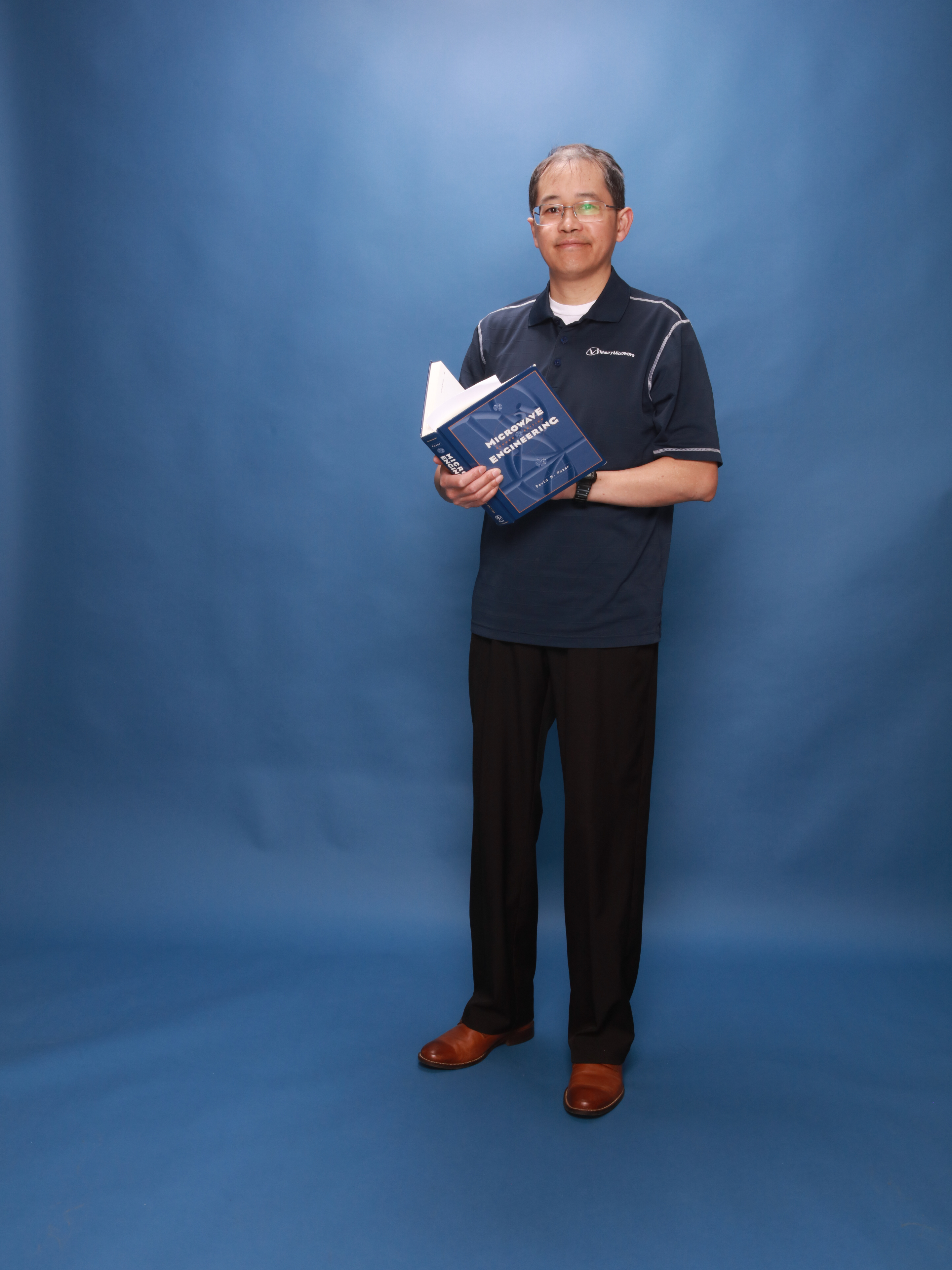 Henry Nguyen standing, wearing a Maury Microwave (engineering firm he previously worked at) shirt, holding an open microwave engineering textbook